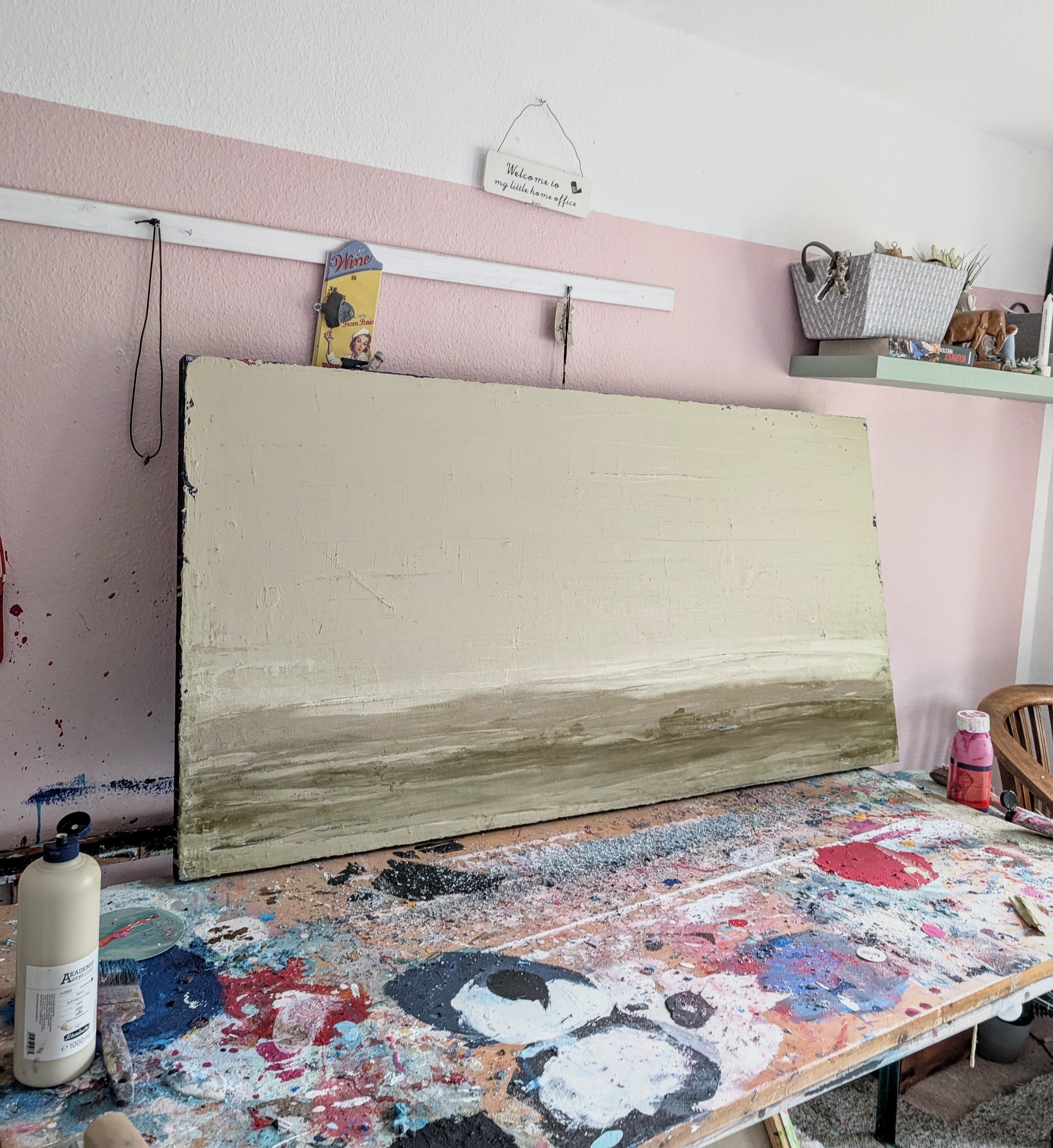 Zwischen Sommer und Herbst II", eine große Unikat-Leinwand der Künstlerin Conny Wachsmann mit abstrakter Spachtelarbeit in neutralen Tönen, liegt auf einem unordentlichen, mit Farbspritzern übersäten Arbeitstisch; Regale und Kunstzubehör säumen die weiß-rosa Wand dahinter.