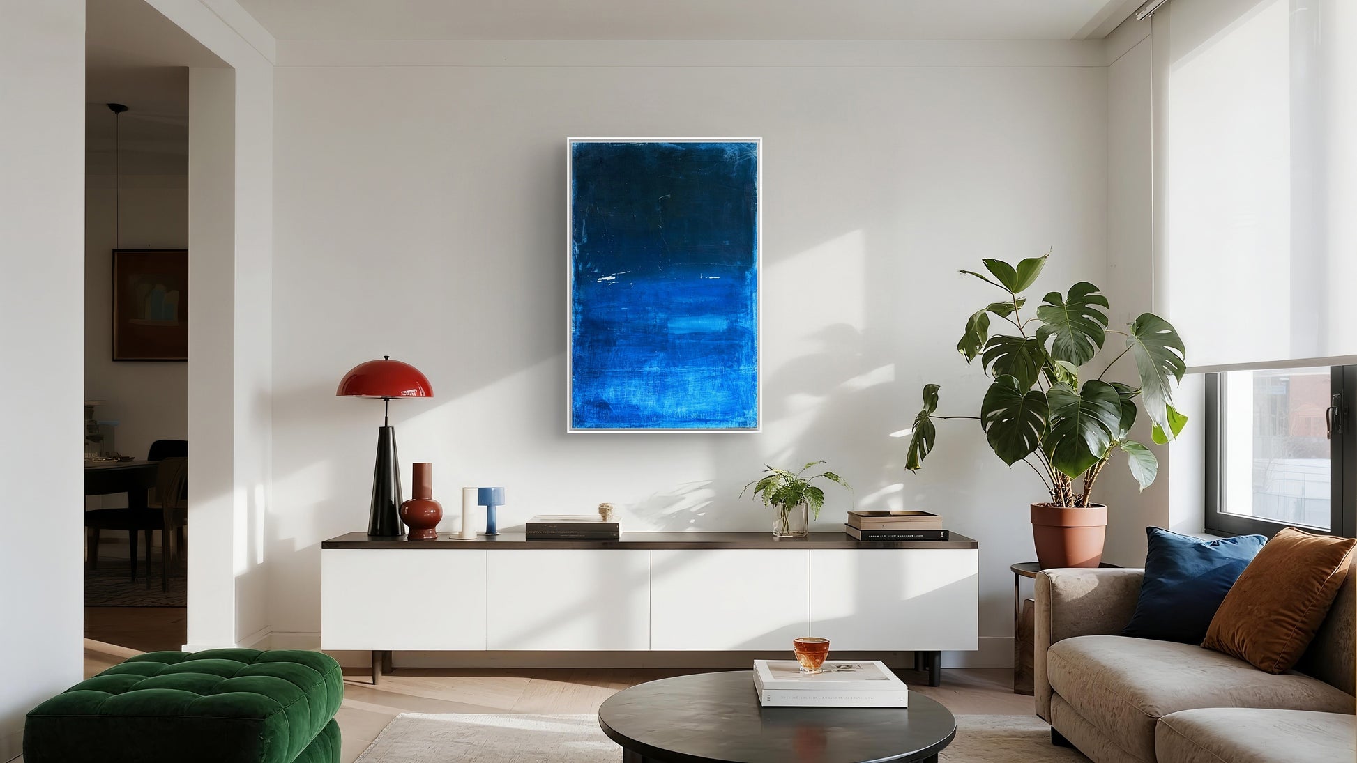Modernes Wohnzimmer mit weißem Sideboard, "Fjord" von Conny Wachsmann an der Wand, grünem Samt-Ottoman, rundem Couchtisch, getöpferter Monstera-Pflanze und natürlichem Licht, das durch ein Fenster fällt.
