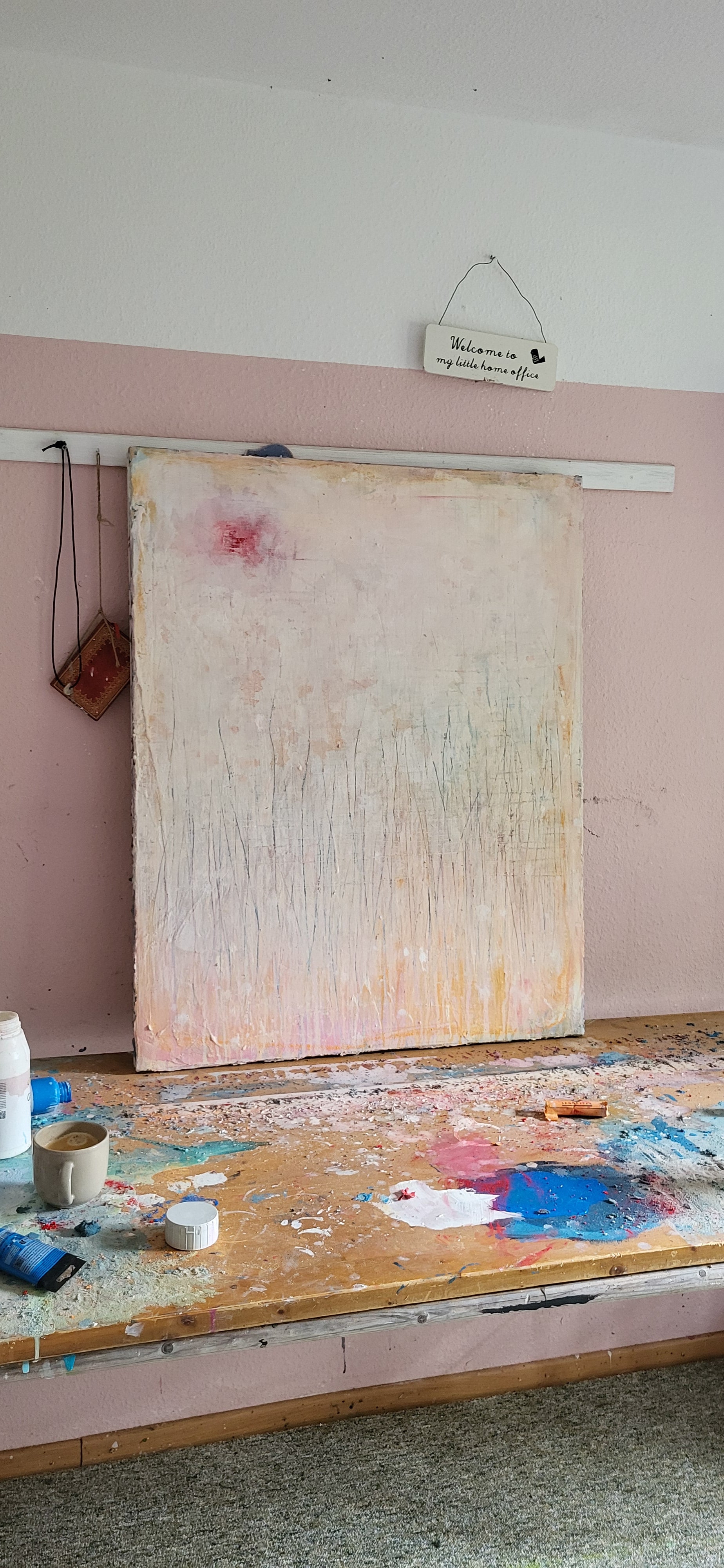 Eine große Leinwand mit dem Titel „Gedankenregen“ der Künstlerin Conny Wachsmann, mit abstrakten Pastelltönen und zarten Linien, lehnt an einer rosa-weißen Wand. Der Tisch darunter ist mit Künstlerbedarf bedeckt. Darüber hängt ein Schild: Willkommen in meinem blauen Kunstcafé – der Heimat einzigartiger Auftragsarbeiten.