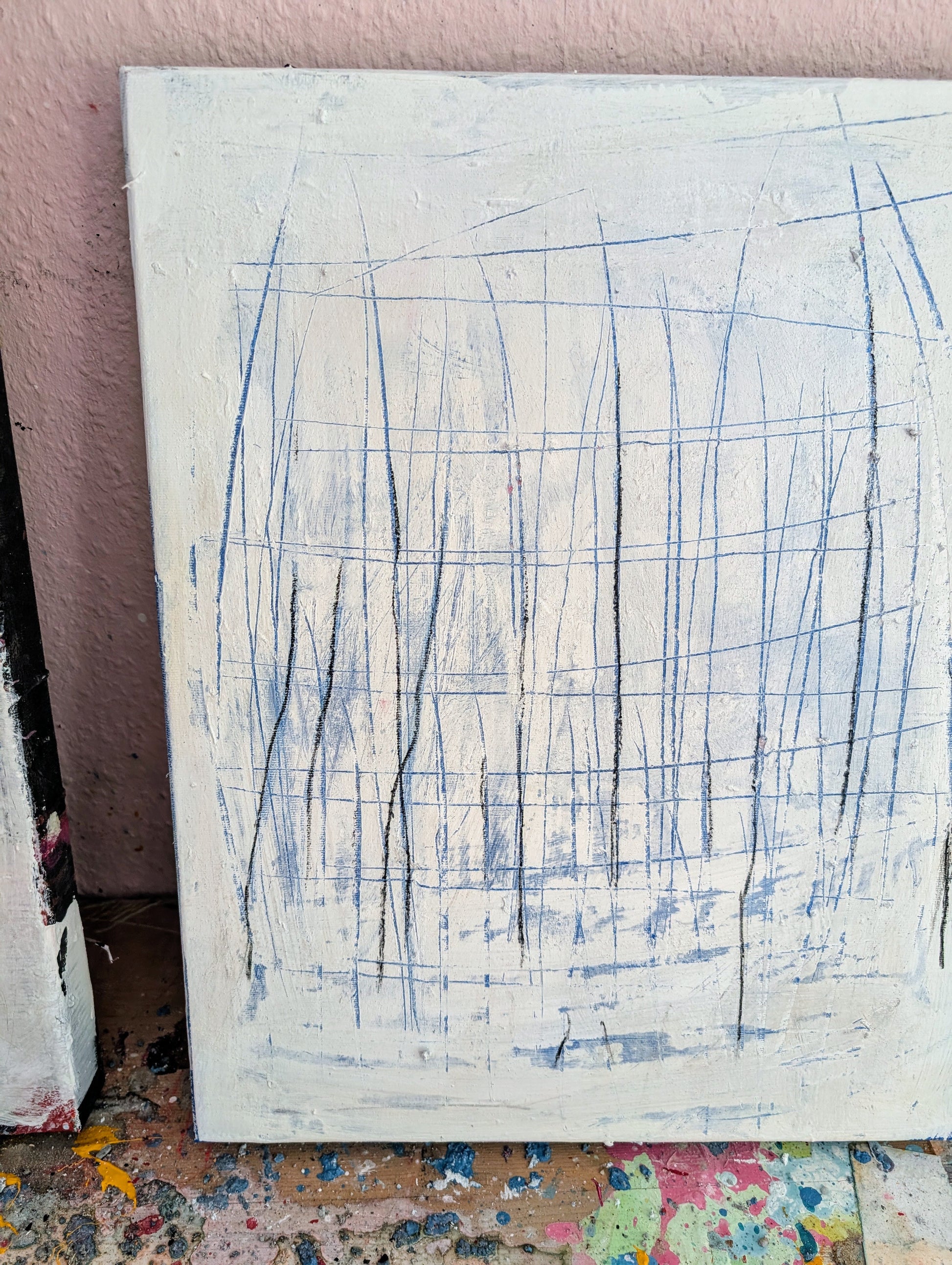 Feldlinien“ von Conny Wachsmann, Künstler: Acryl auf Leinwand als abstraktes Unikat mit feinen Linien in Schwarz & Blau auf weißer, texturierter Fläche, angelehnt an eine rosa Wand. Farbkleckse zieren die Holzoberfläche darunter.