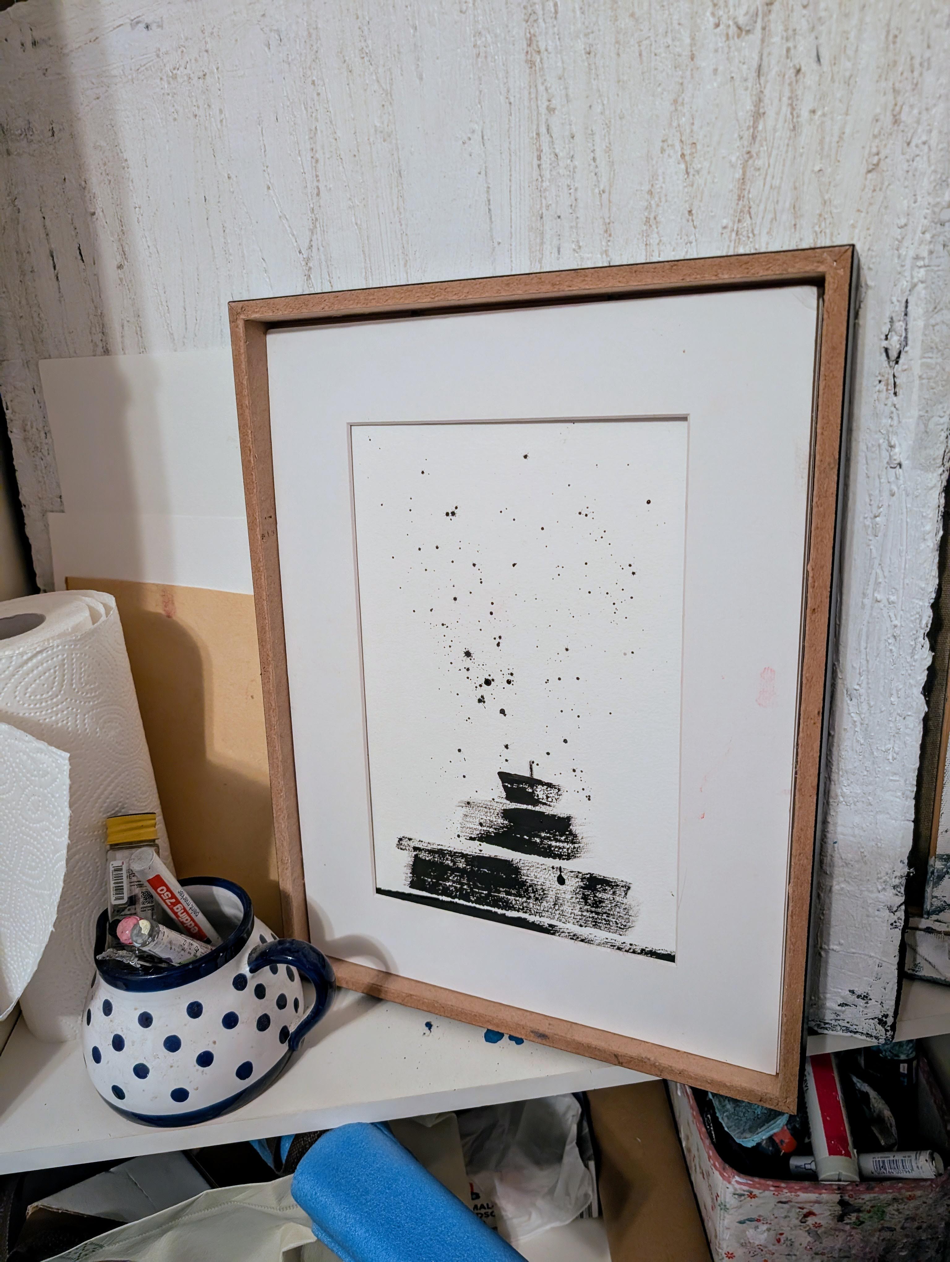 Eine gerahmte "Zeichnungen & Reduktion V" von Conny Wachsmann lehnt an einer strukturierten weißen Wand. Daneben steht ein blau gepunkteter Keramikkrug mit Kunstzubehör, der diesen kreativen, minimalistisch inspirierten Raum vervollständigt.